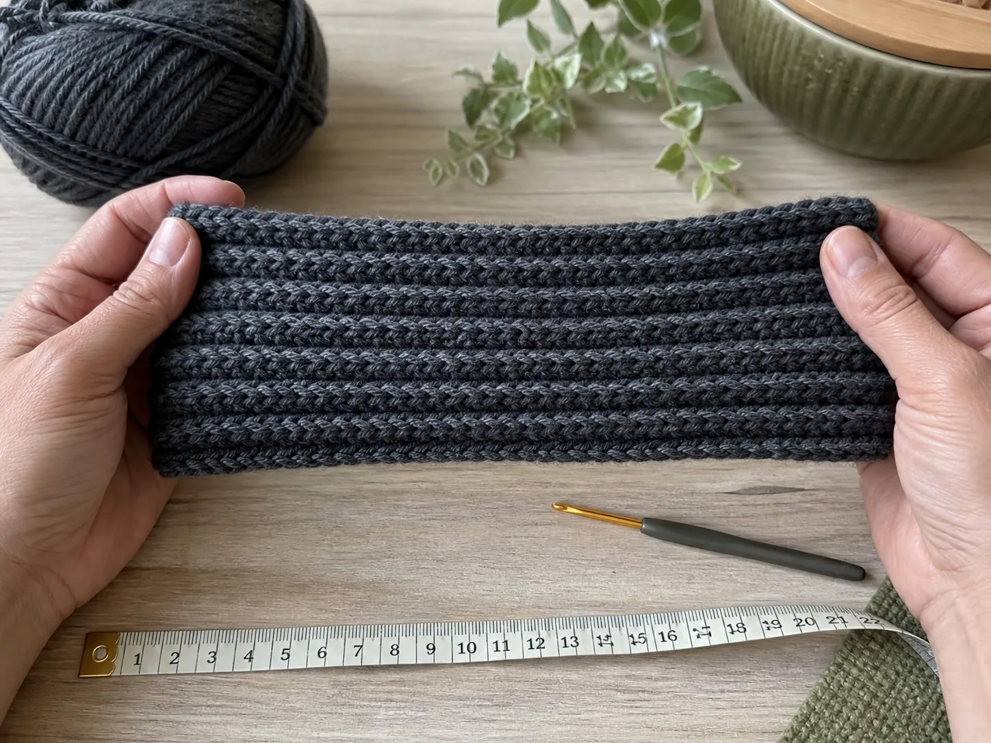 Hands holding a charcoal ribbed crochet band swatch beside a measuring tape on a wood table.