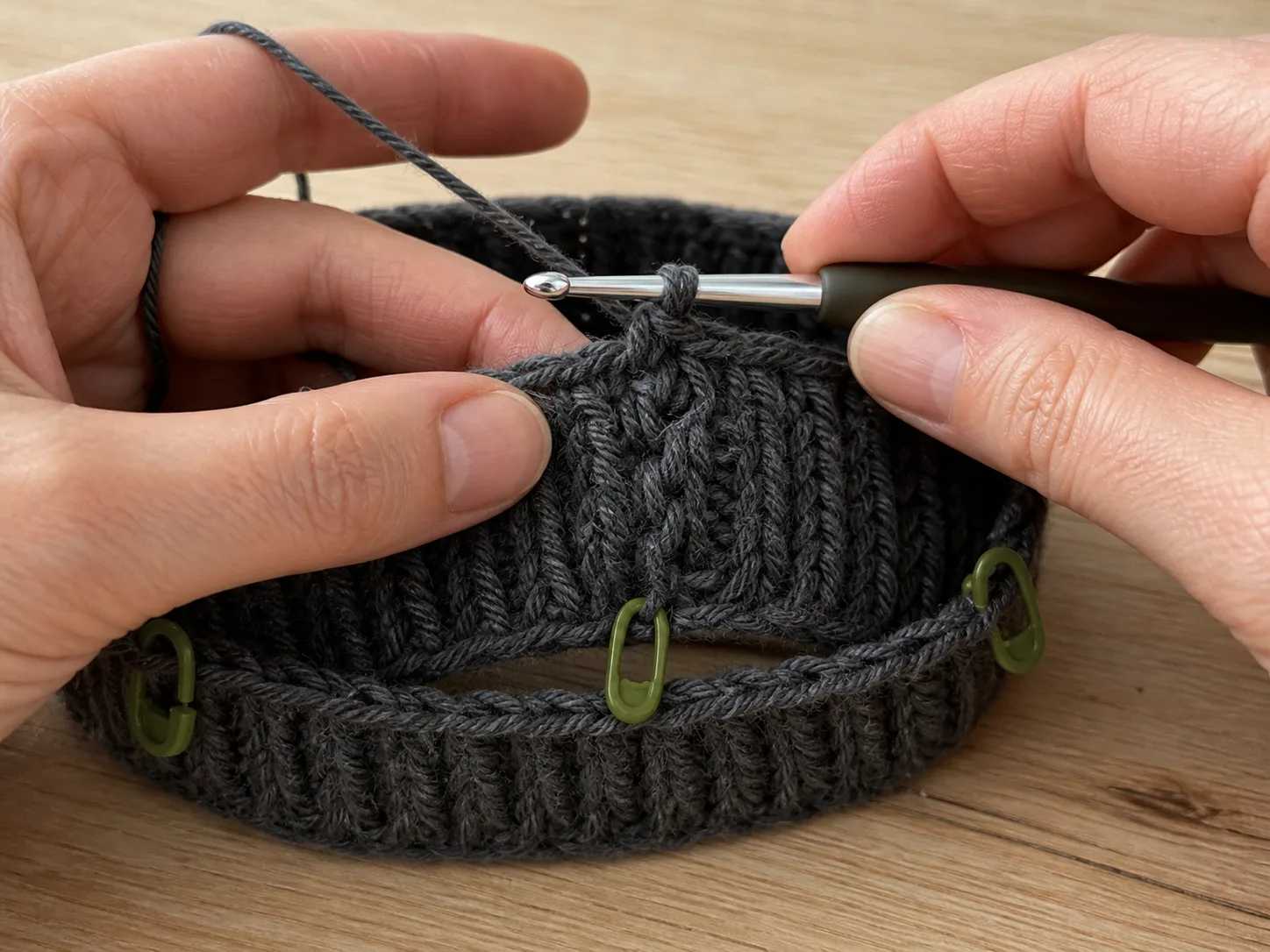 Hands crocheting the first body round into a joined charcoal ribbed band marked with olive stitch markers.