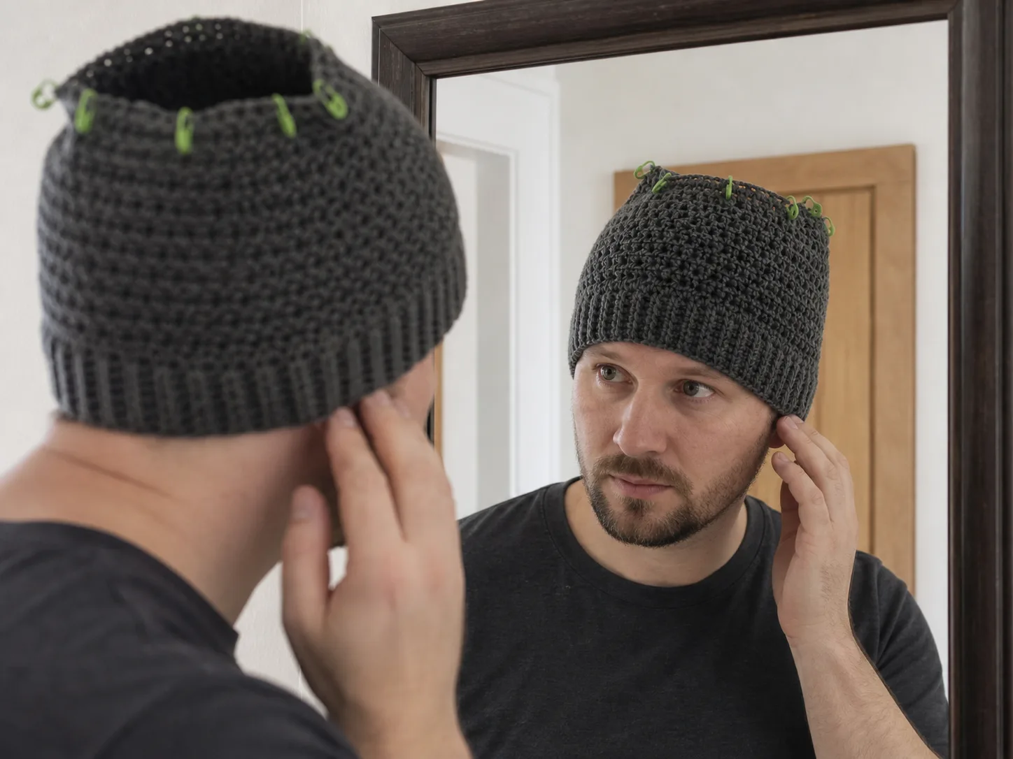 Adult man checking ear coverage in a mirror while wearing an unfinished open-top charcoal crochet beanie with stitch markers.
