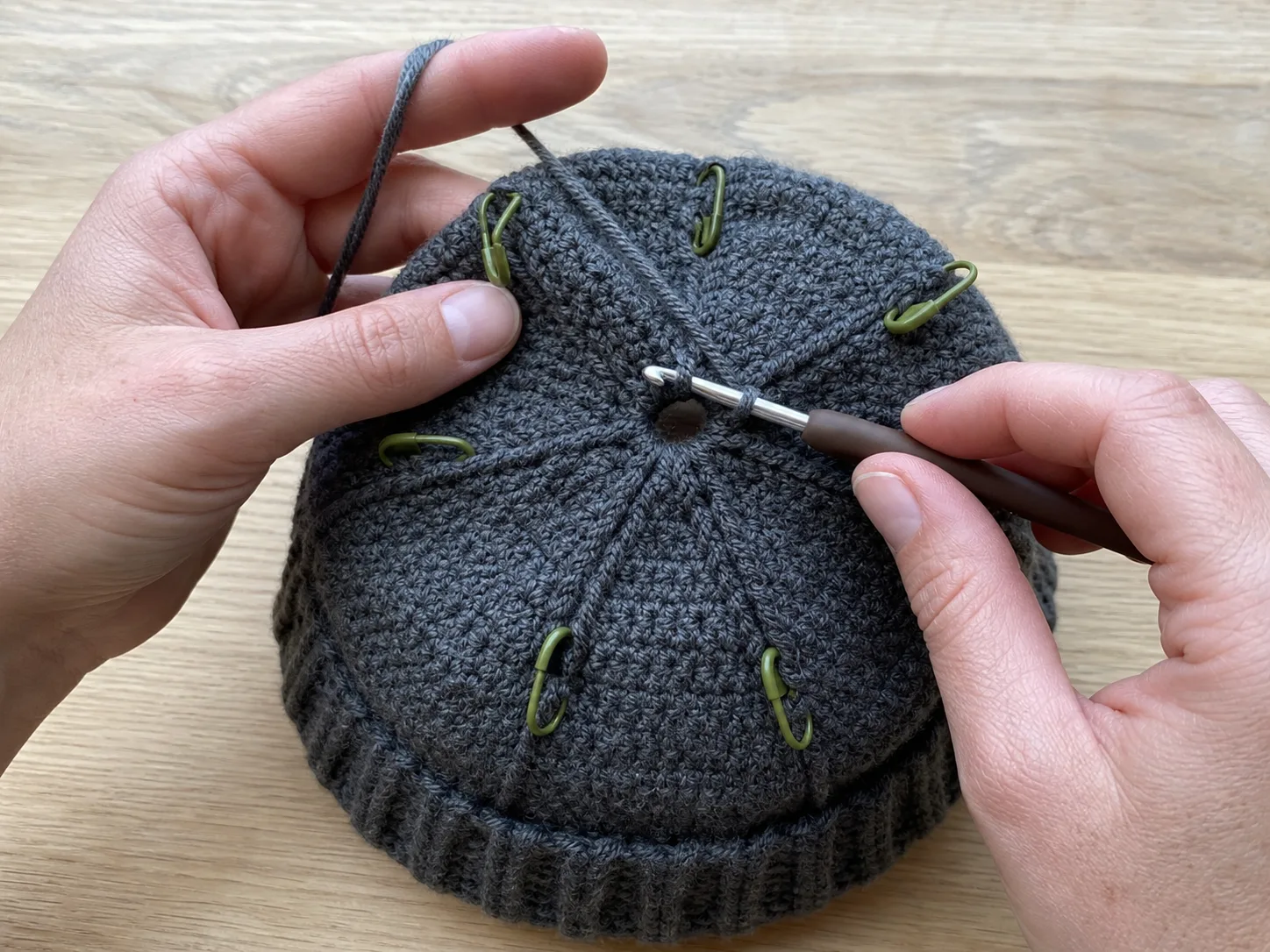 Hands decreasing a bottom-up charcoal crochet beanie crown toward a small center opening with olive stitch markers dividing the sectors.
