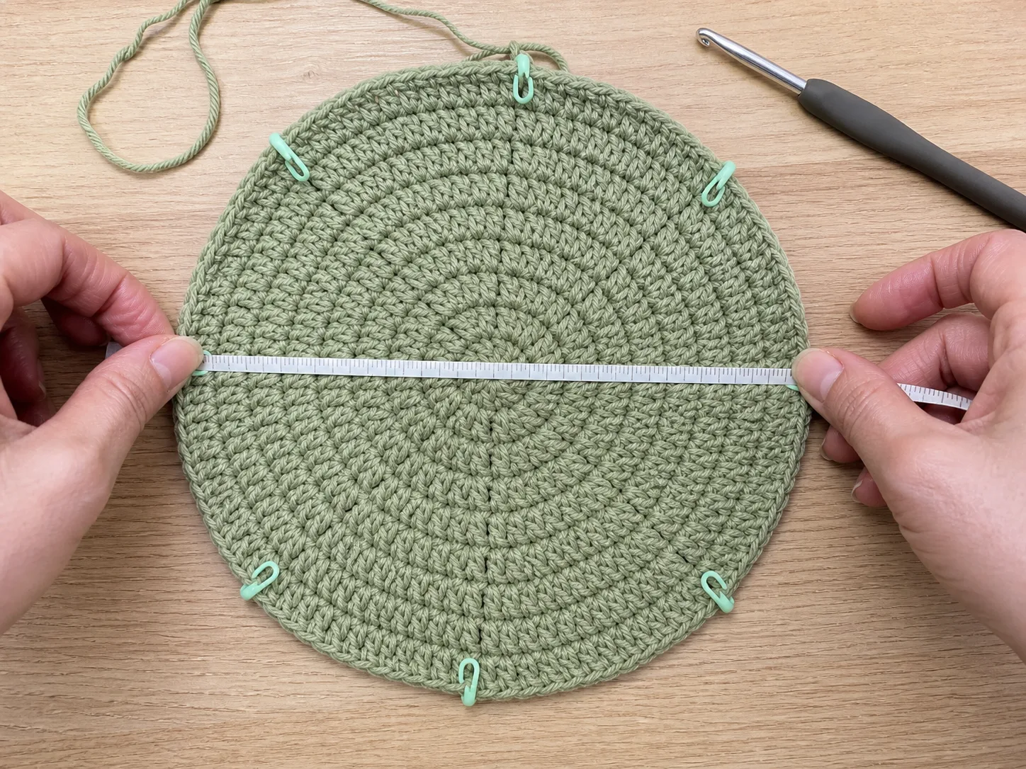 Hands positioning a measuring tape across a flat sage crochet crown with stitch markers.