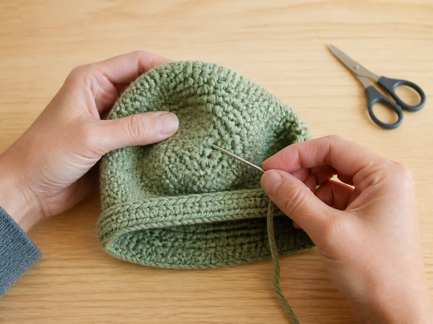 Hands weaving a yarn tail inside a sage crochet baby beanie with a blunt yarn needle.