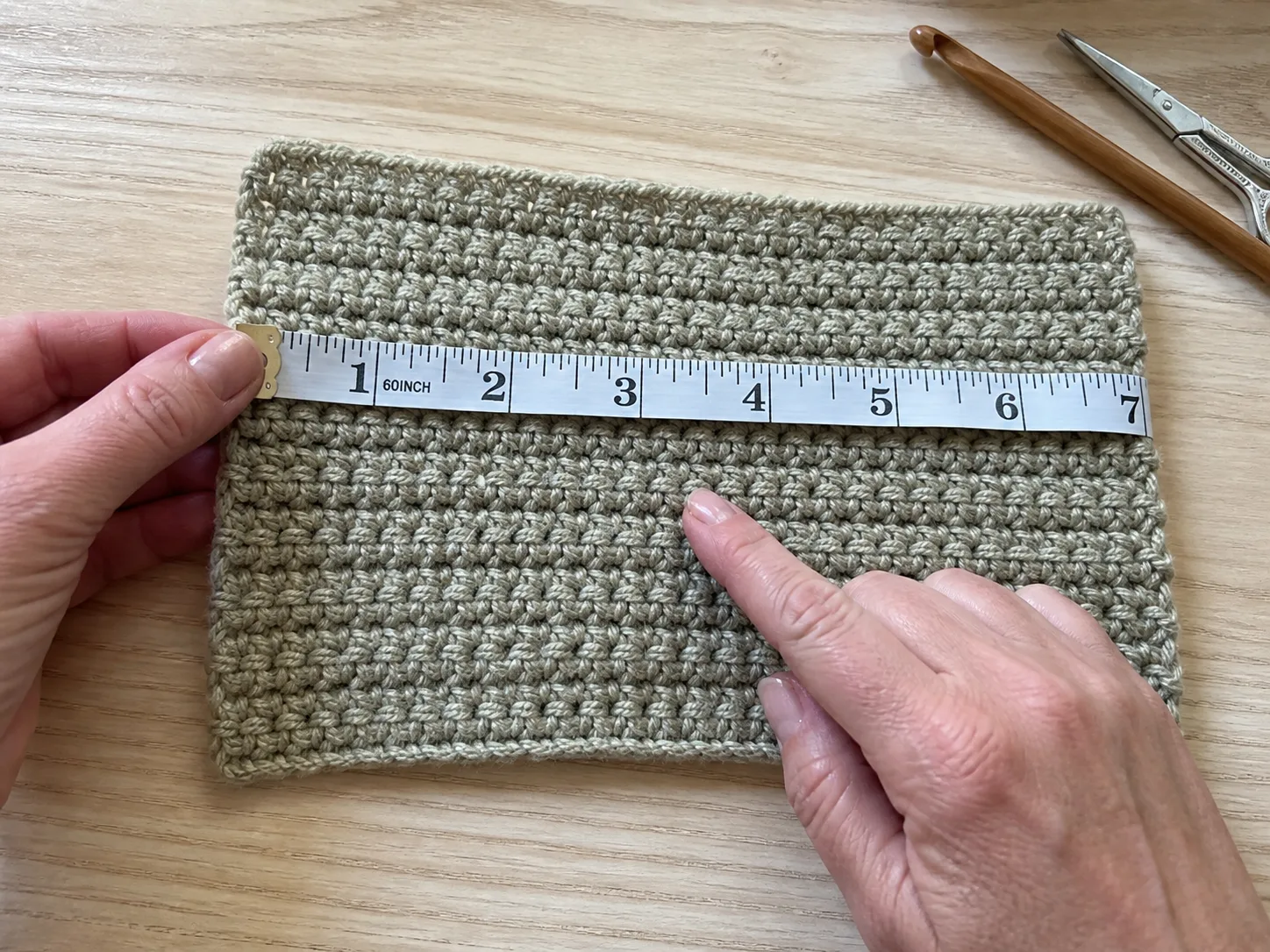 Adult hands measuring the center of an oatmeal-sage half-double-crochet swatch with a flexible tape on a wood table.