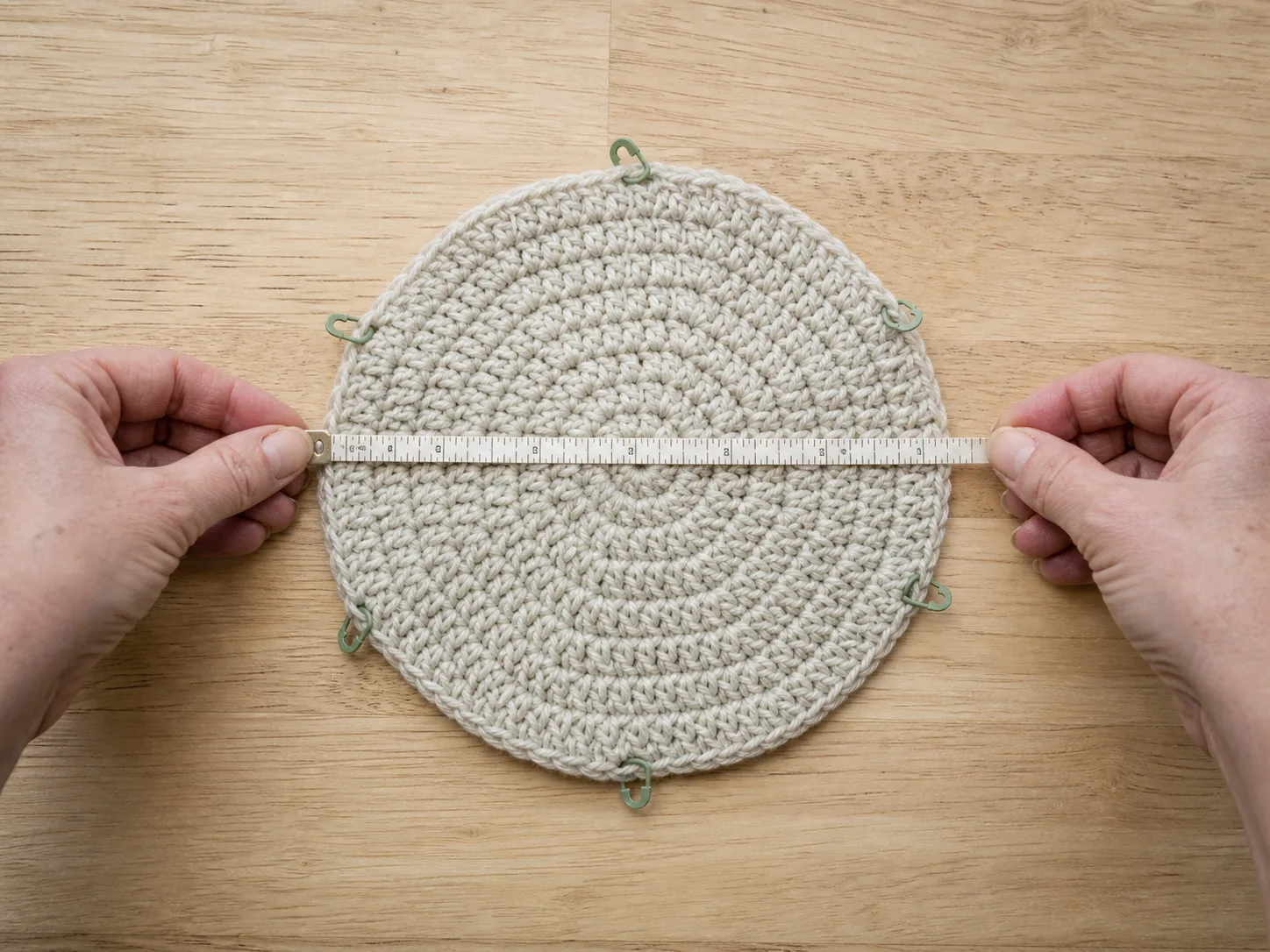 Adult hands measuring a small flat oatmeal crochet crown across its center with stitch markers around the edge.