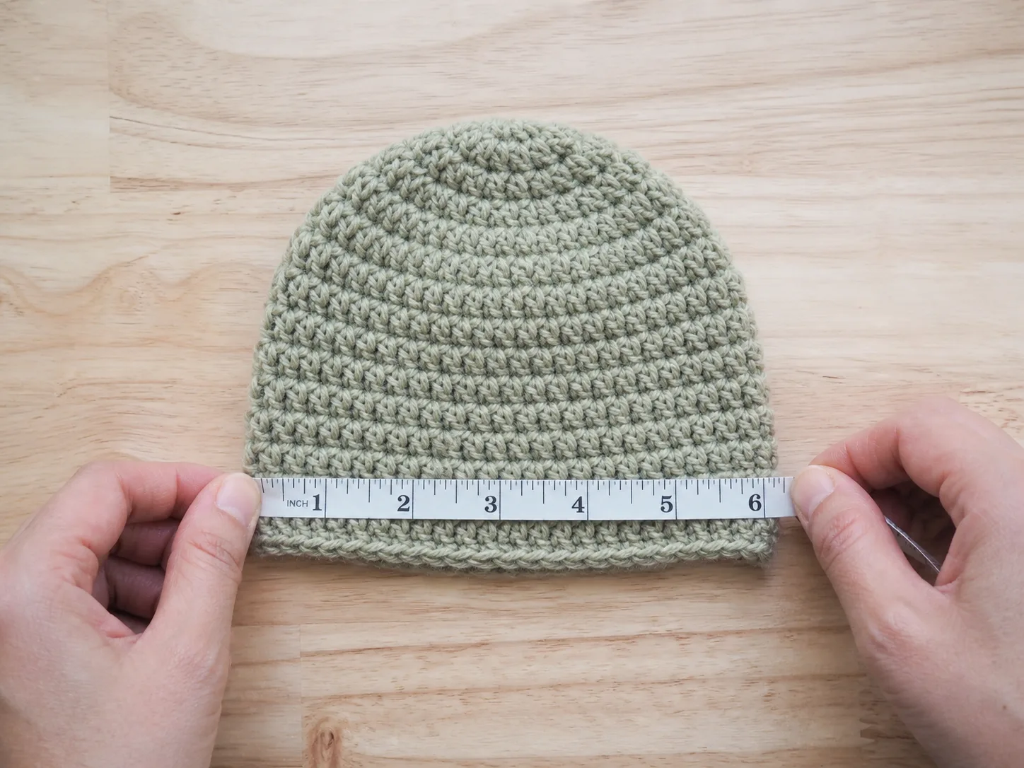 Adult hands measuring the relaxed flat width of a plain short newborn crochet beanie on a wood table.