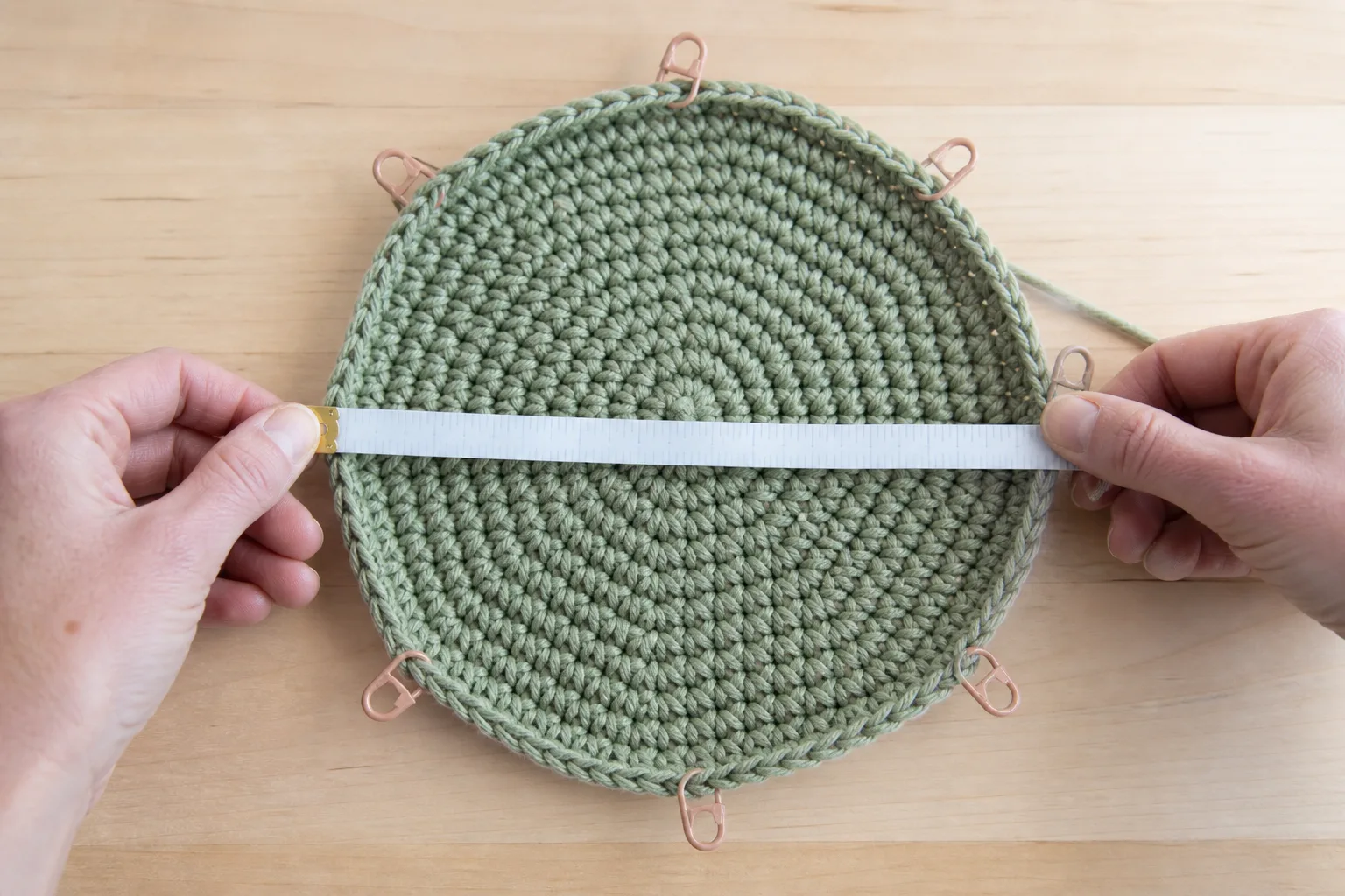 Hands measuring straight across a small flat sage-green crochet crown with eight stitch markers around the edge.