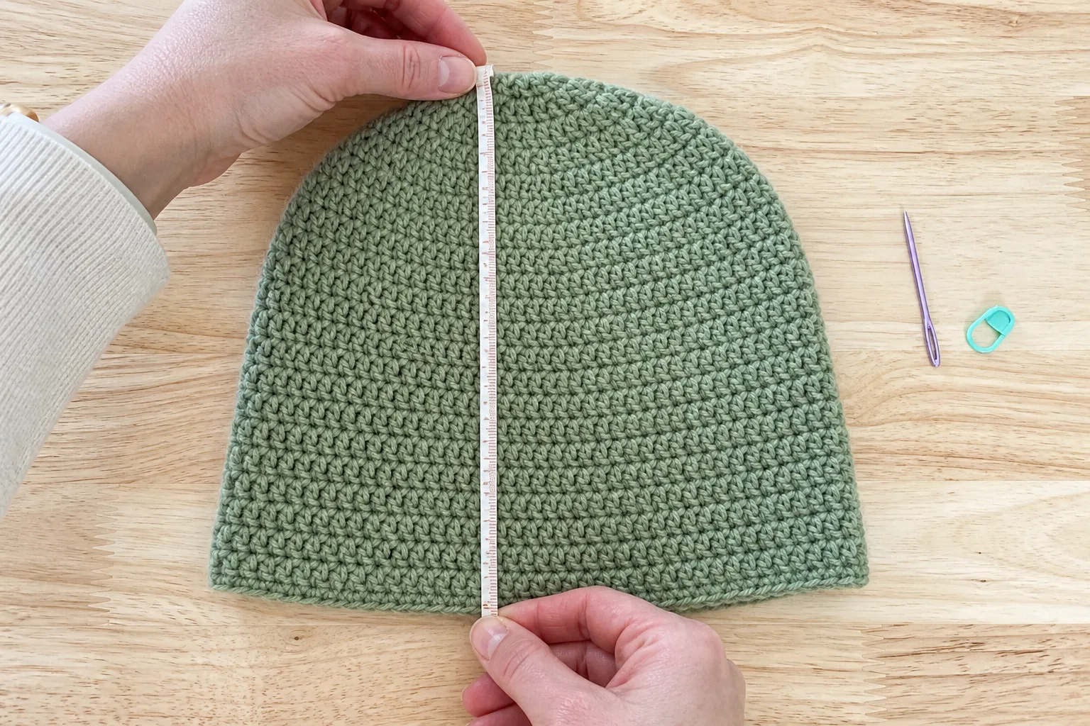 Adult hands measuring a finished plain sage-green child beanie from the crown center to the fully unfolded lower edge on a wood table.