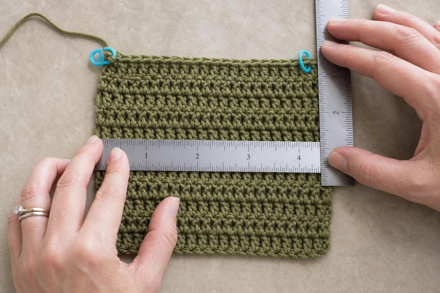 Adult hands measuring stitch width and row height on an olive half-double-crochet swatch with rulers and stitch markers.