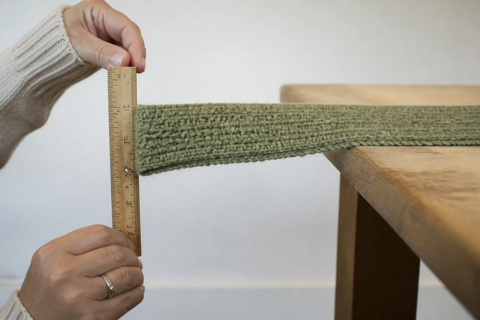 Adult hands measuring an olive crochet swatch strip projecting from a wooden table while its unsupported tip bends downward.