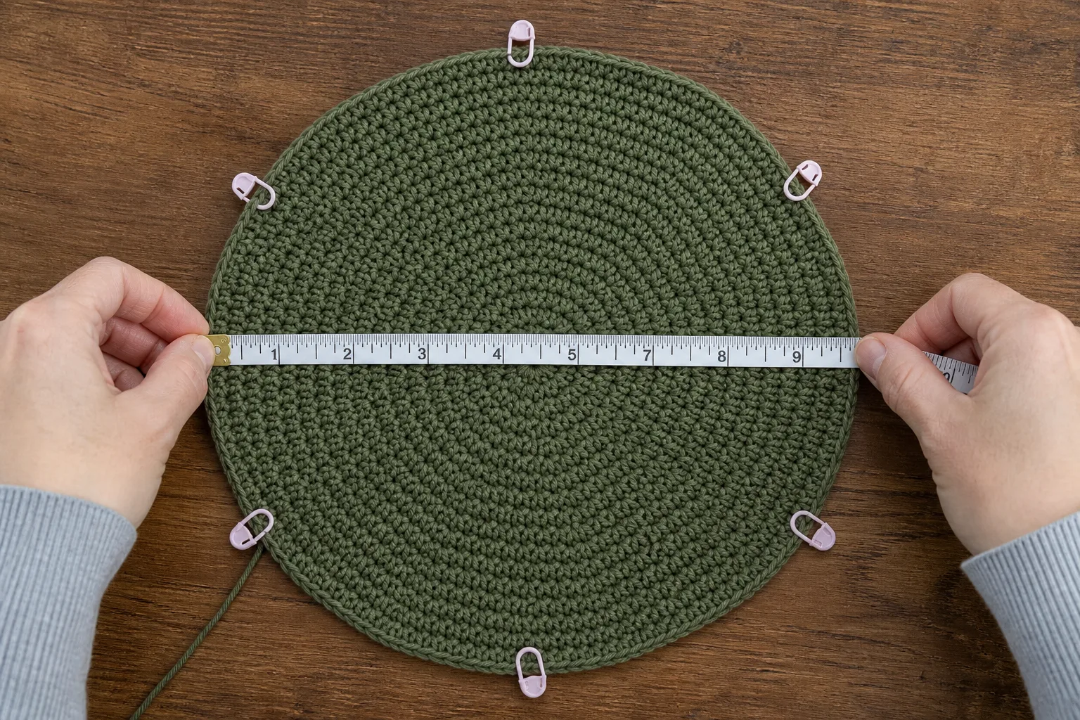 Adult hands measuring the diameter of a flat olive crochet crown with removable markers spaced around its perimeter.