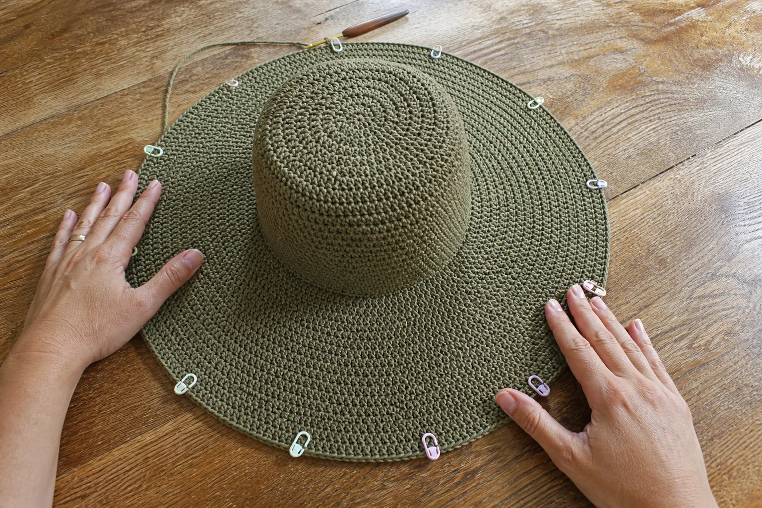 Adult hands checking a broad flat olive crochet sun-hat brim with stitch markers distributed around the outer round.