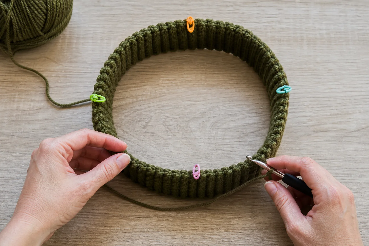 Adult hands preparing to crochet body pickups around an olive ribbed cuff ring marked into four equal quarters.