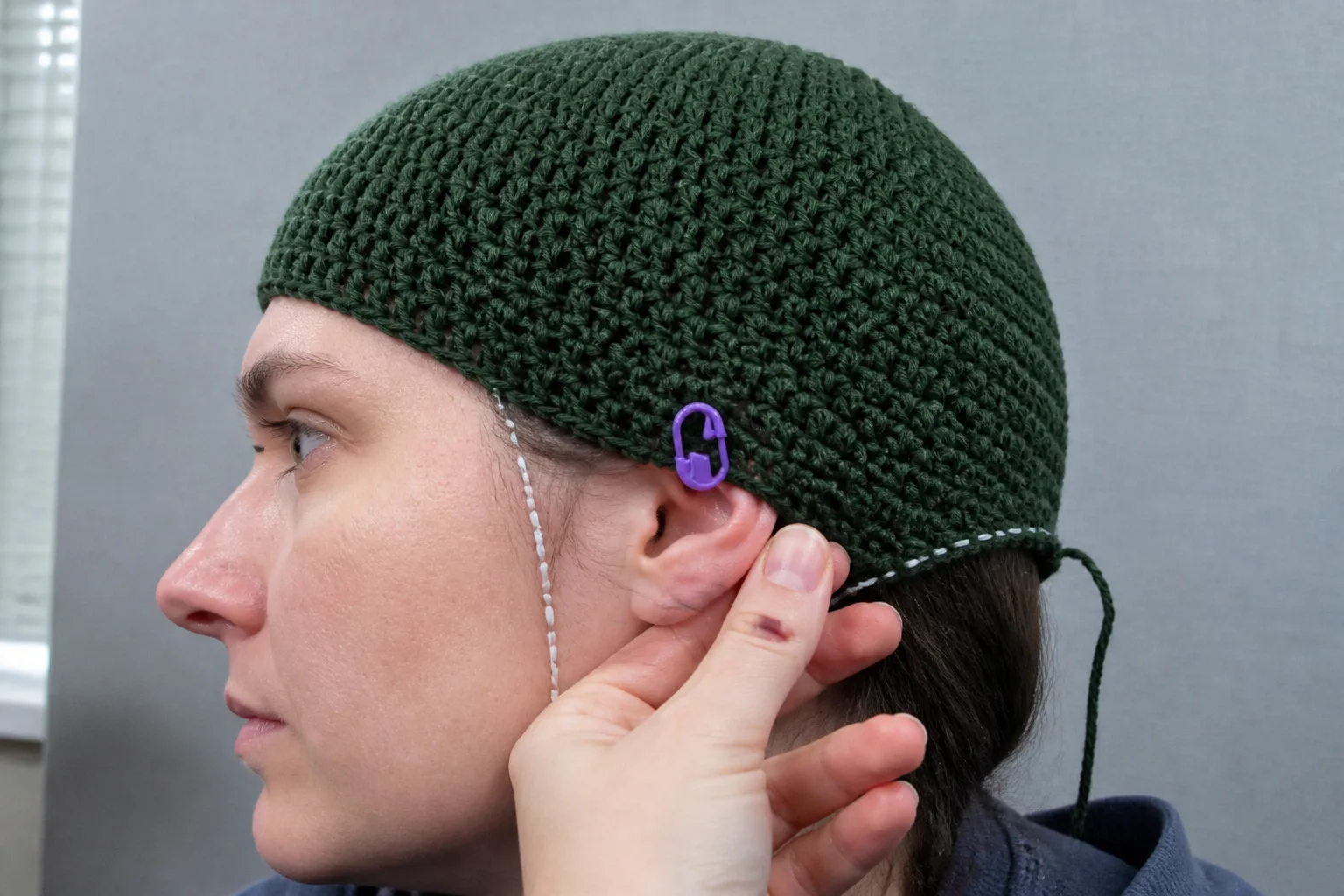 Side-profile close-up of an adult wearing an unfinished green beanie with a marker aligned directly above the ear center.