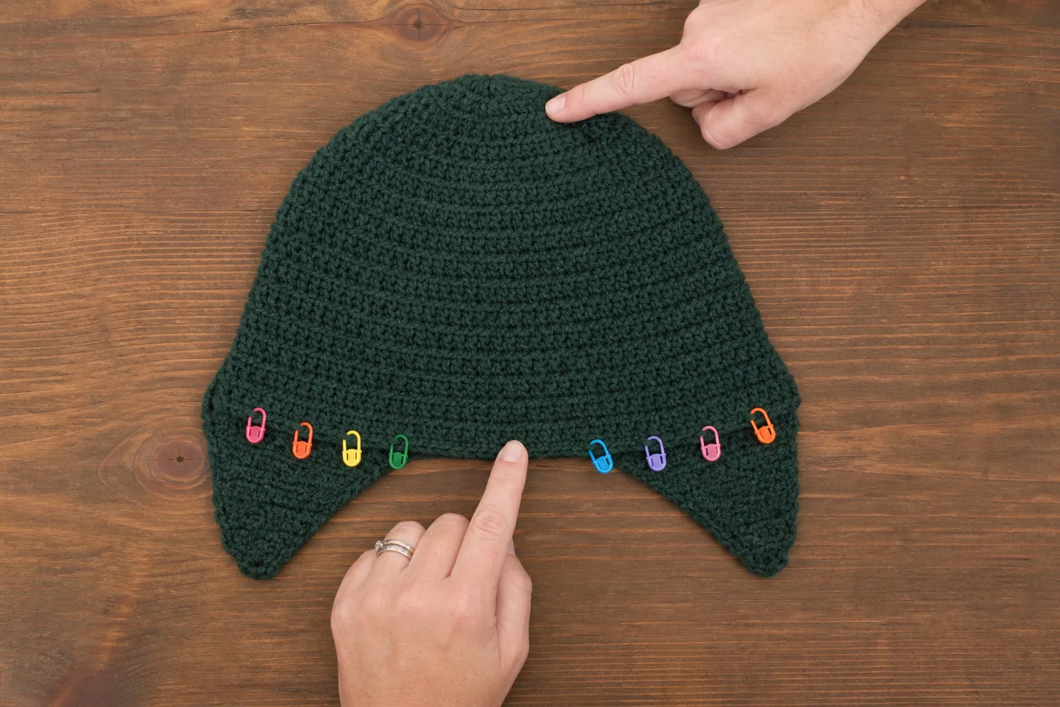 Overhead view of a forest-green crochet hat with two tapered ear areas and colorful markers while hands indicate its center landmarks.