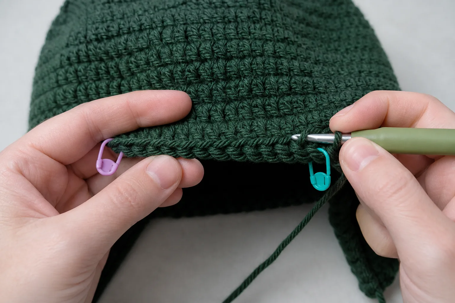 Macro view of adult hands crocheting across a marked section of the lower edge of a forest-green beanie.