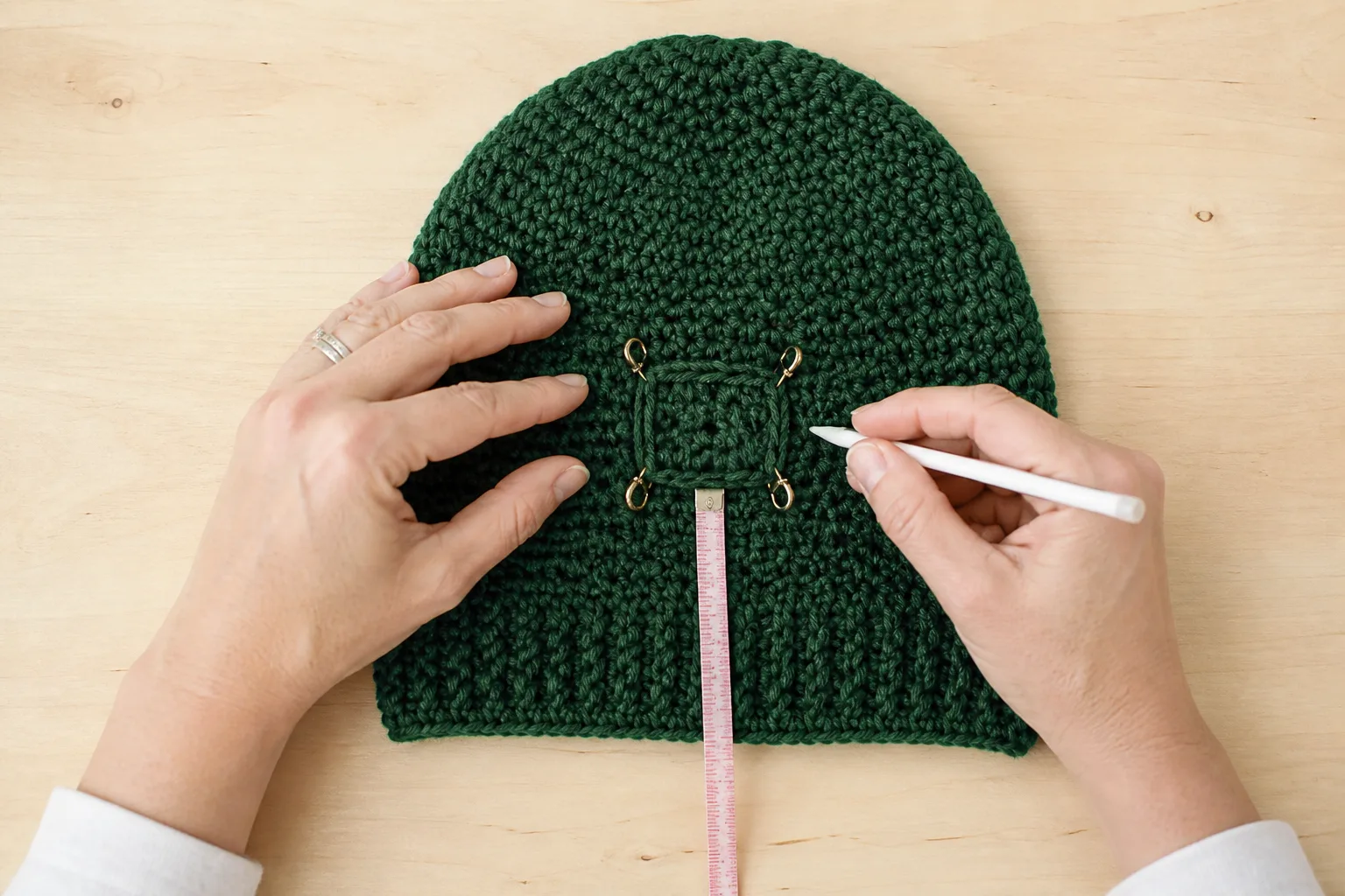 Adult hands marking a rectangular ponytail window with brass stitch markers on a forest-green crochet beanie.