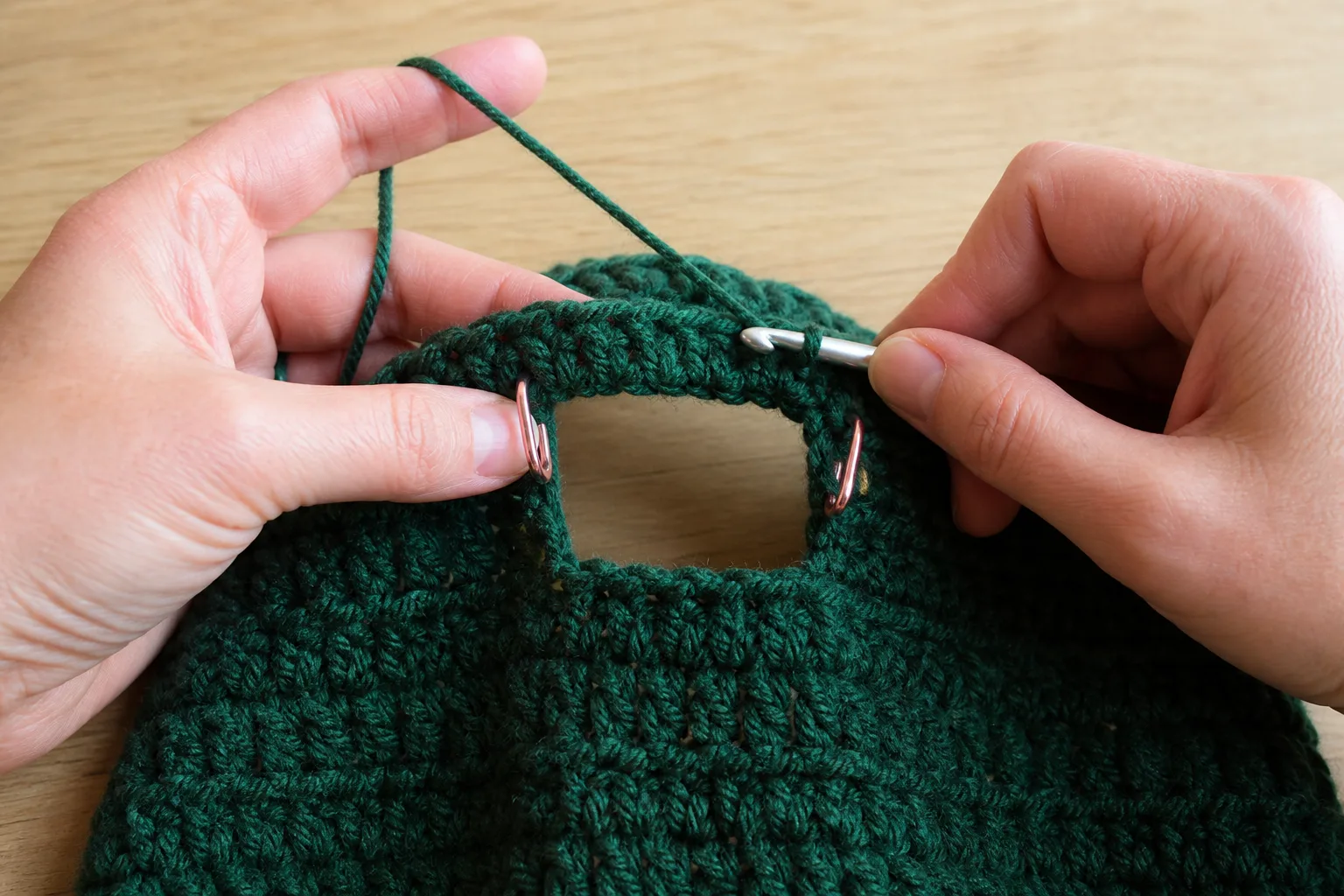 Close view of adult hands crocheting around a marked opening in a forest-green crochet hat.