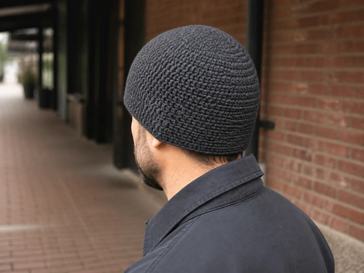 Adult man seen from a rear three-quarter angle wearing a fitted charcoal crochet beanie with a smooth crown.