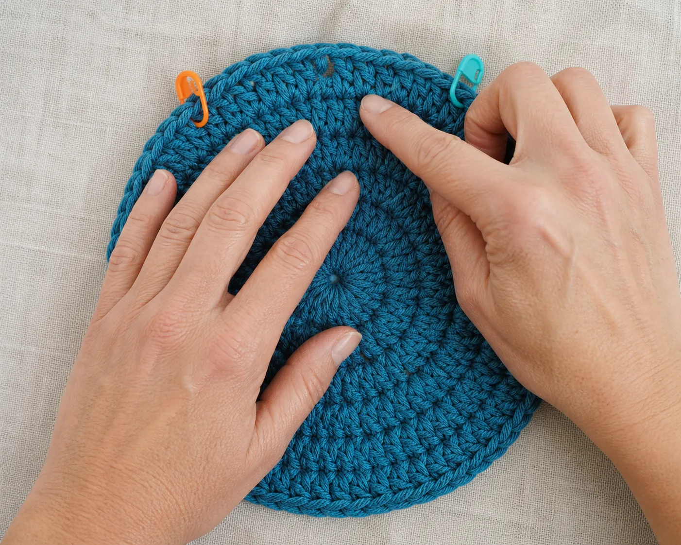 Adult hands count V-shaped stitches around a teal crochet crown between orange and blue markers.