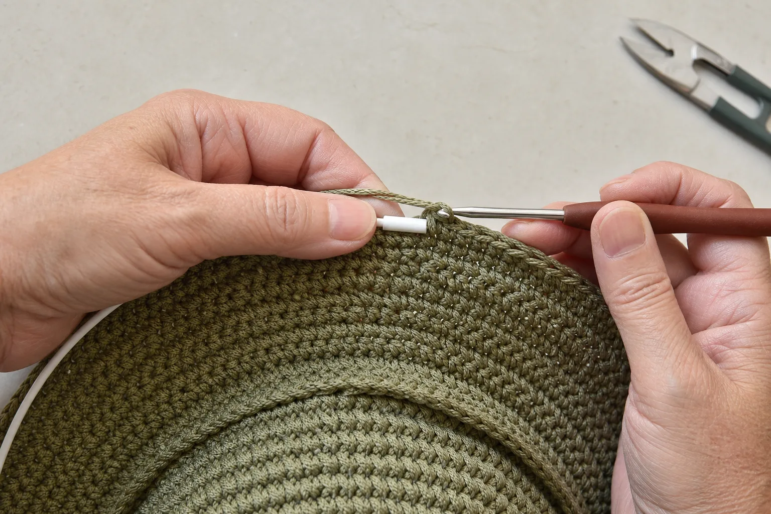 Hands crochet over flexible white millinery support at the outer edge of an olive hat