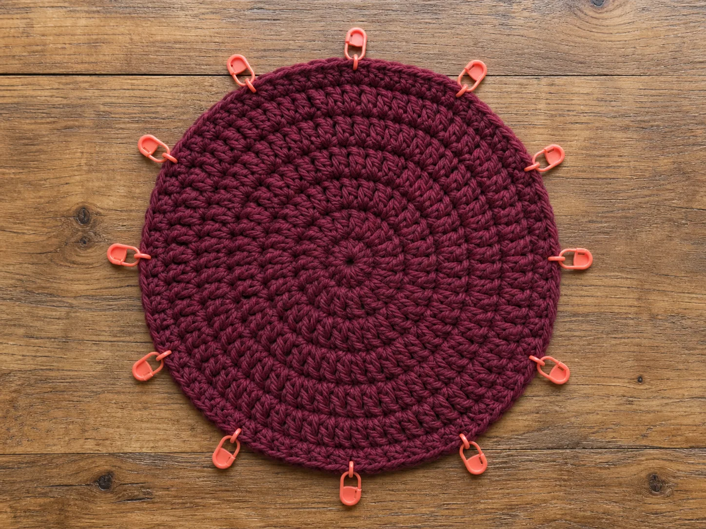 Twelve coral stitch markers divide a burgundy crochet disc into equal sectors