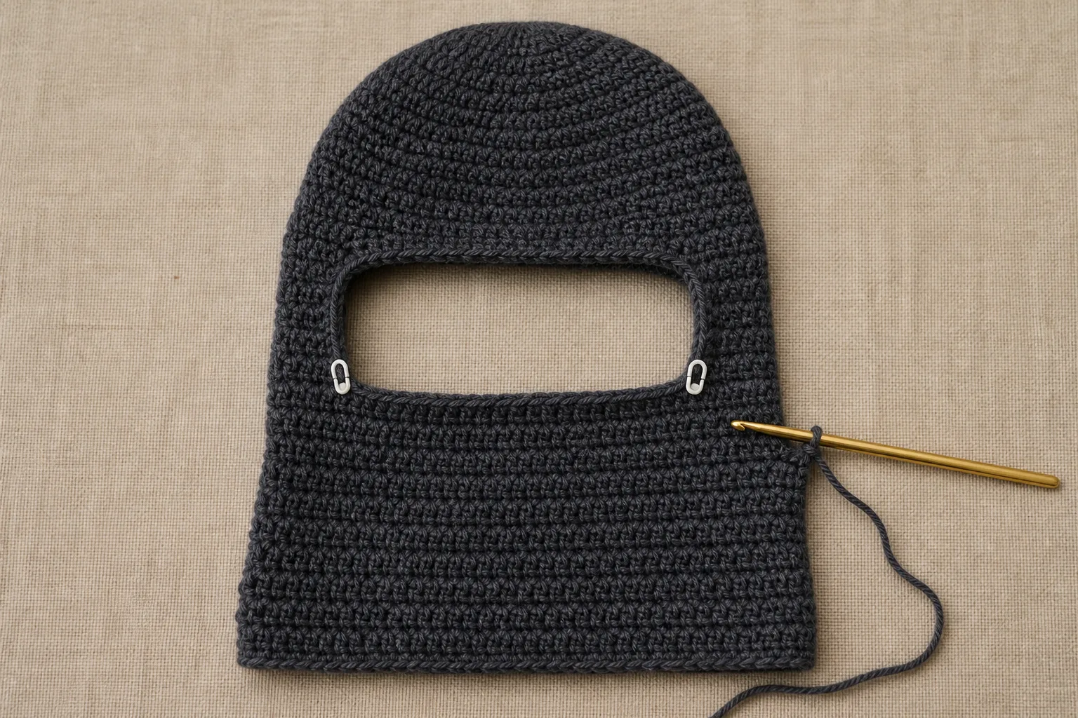 An in-progress charcoal crochet balaclava lies flat with a centered face gap and two white edge markers